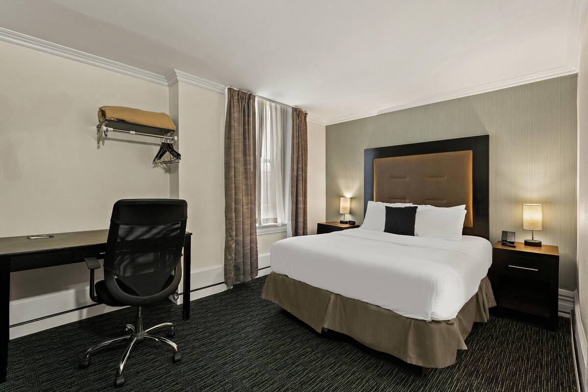 A hotel room with a bed, a desk, a chair, a nightstand with a lamp, and a window with curtains.