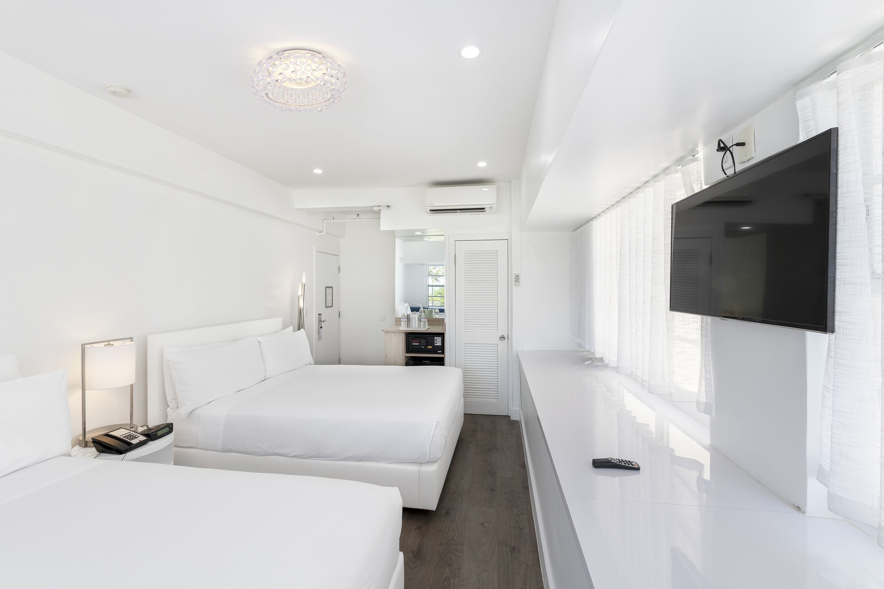 Et moderne hotelværelse med to senge, et fladskærms-tv, et natbord med lampe og udsigt til en gang med en dør.