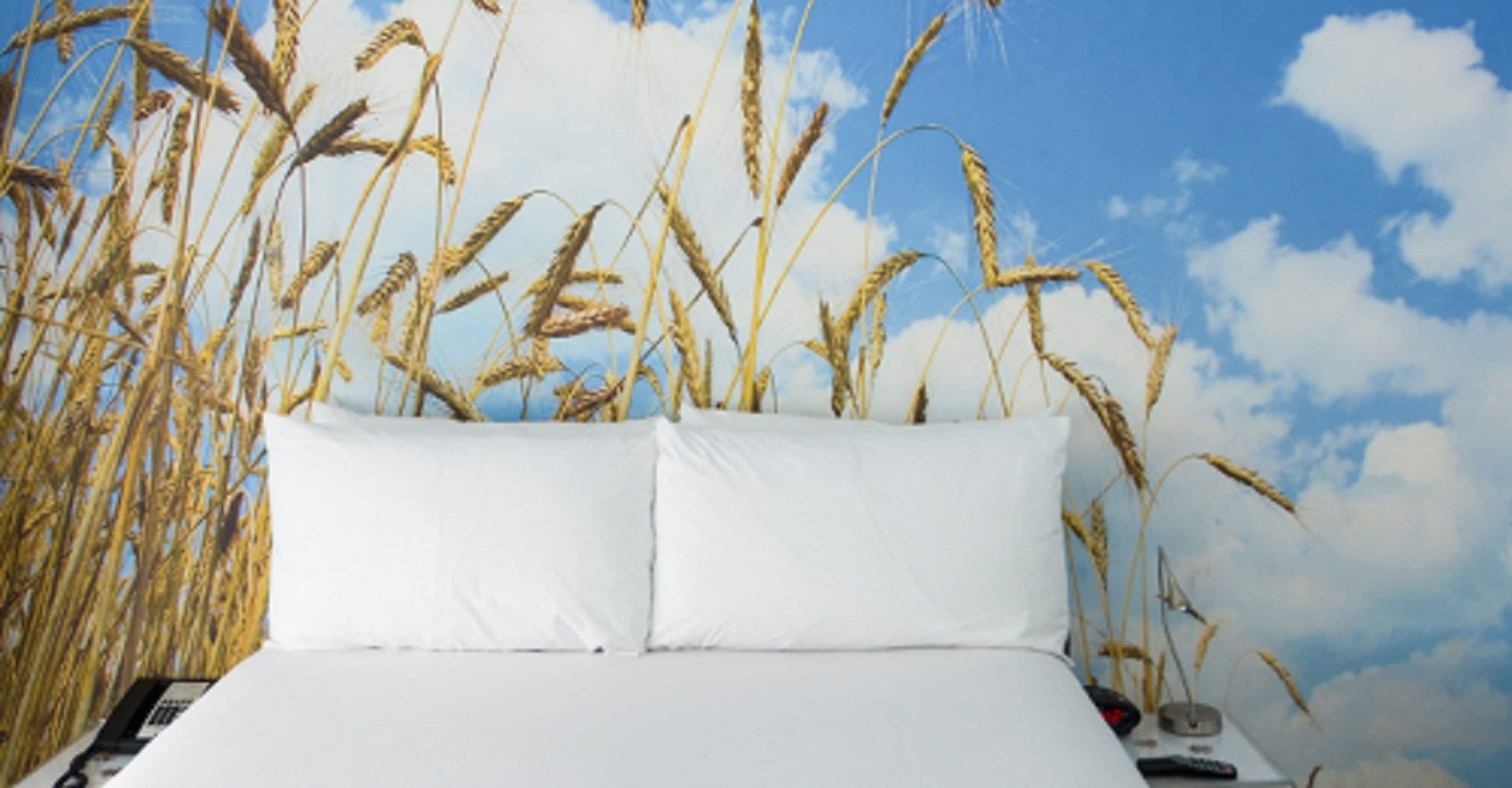Una cama con ropa blanca frente a un fondo de espigas de trigo y un cielo azul con nubes.