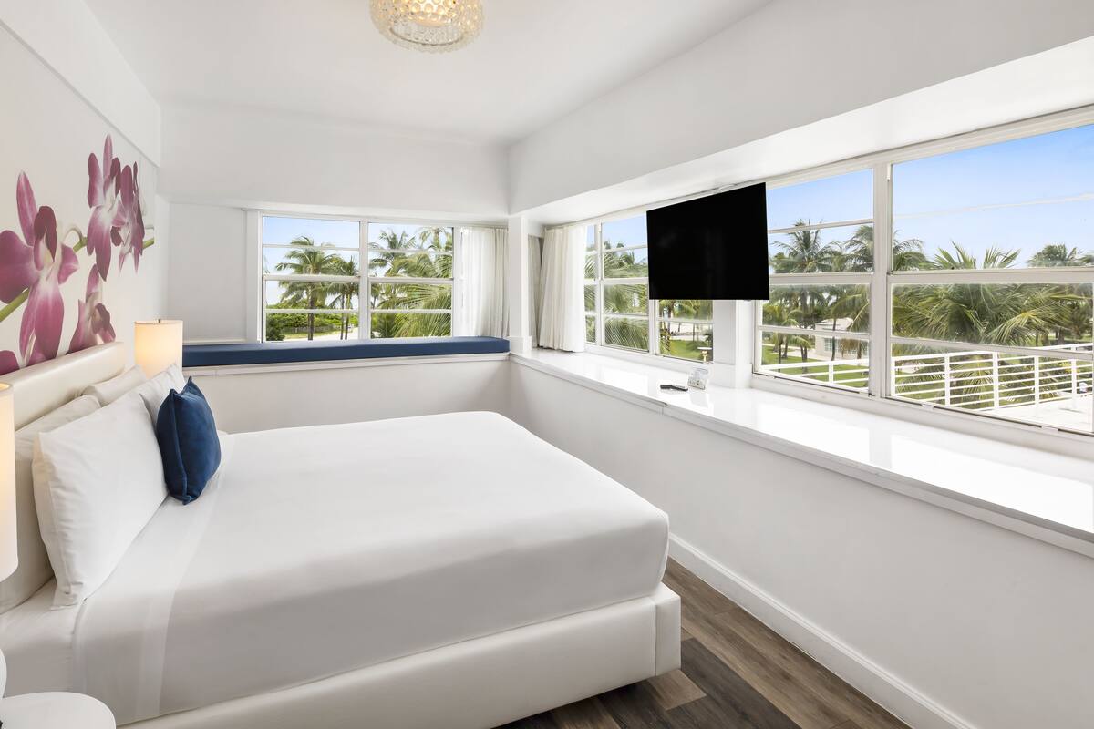 A modern hotel room with a large bed, a TV, and a view of palm trees.