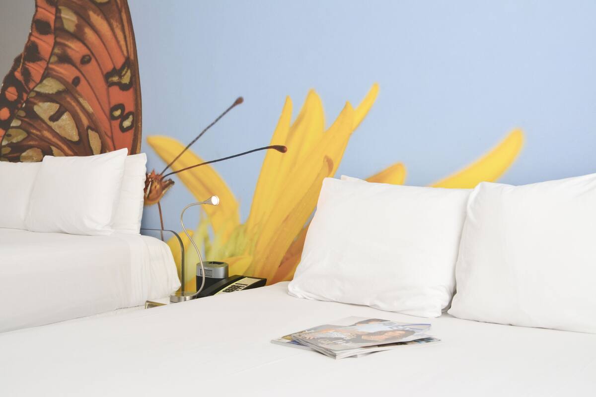 A hotel room with a bed, white pillows, a magazine on the bed, a lamp, and a wall with a large yellow flower painting.