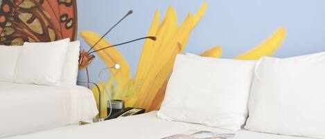 Habitación de hotel con cama, almohadas blancas, una revista sobre la cama, una lámpara y un cuadro de una flor amarilla grande en la pared.