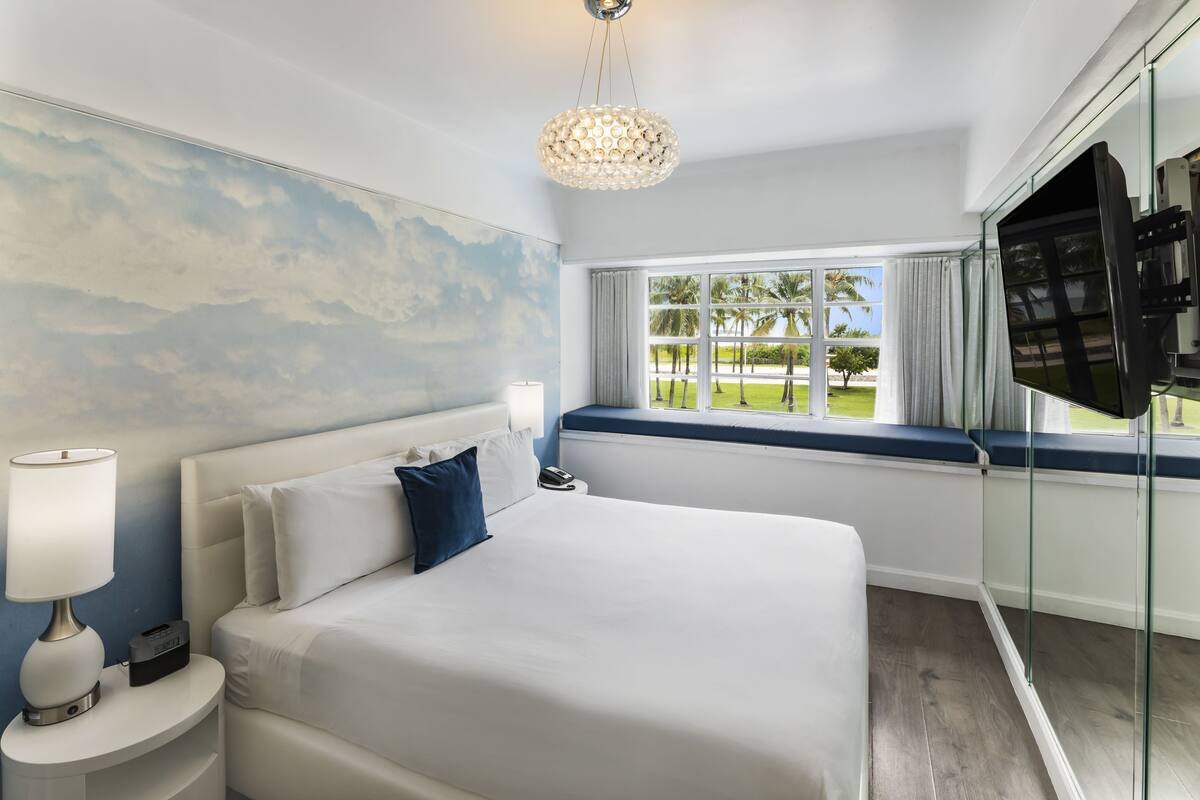 A modern hotel room with a large bed, a nightstand with a lamp, a glass wall with a TV, and a window with a view of palm trees.