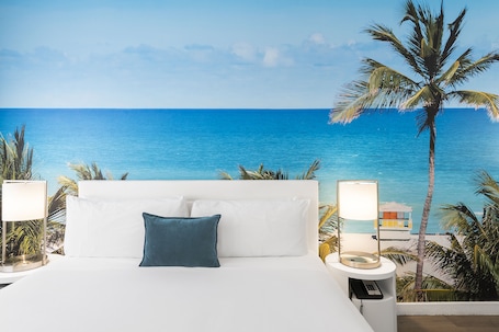 A bed with white linens and a blue pillow, flanked by two bedside lamps, set against a backdrop of a clear blue sea and palm trees.