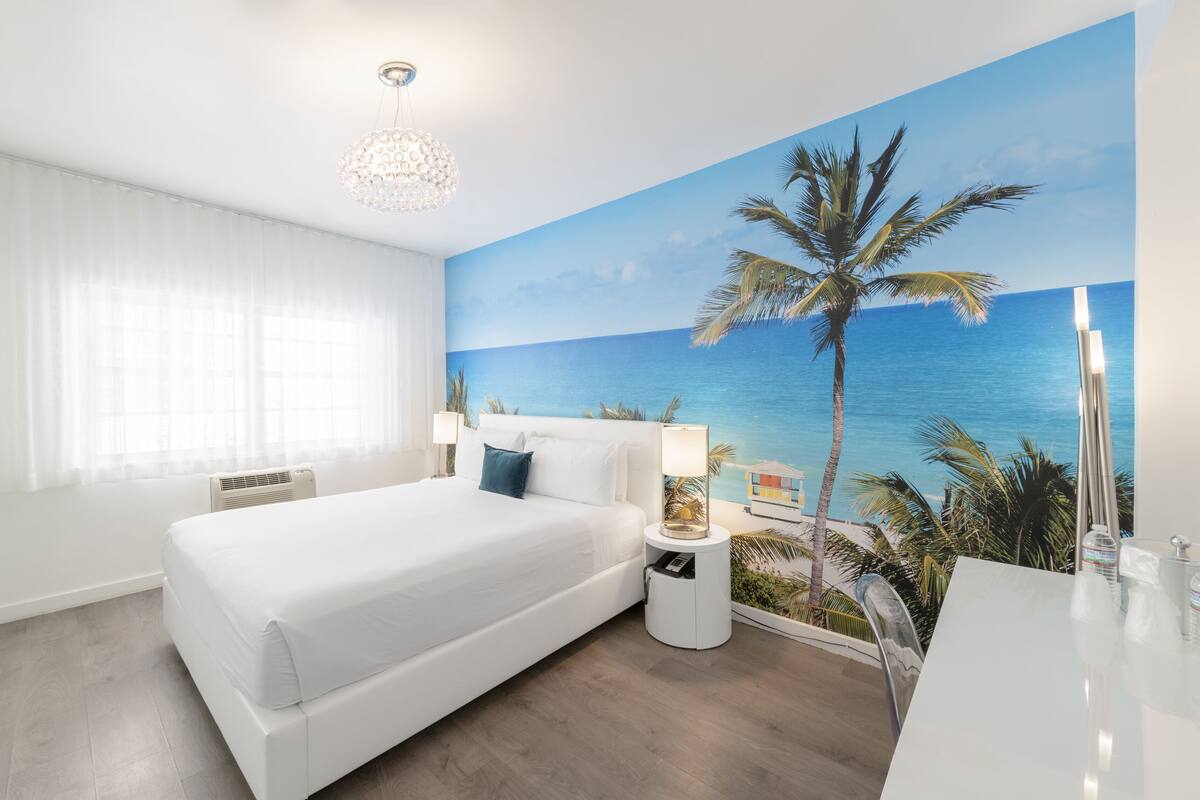 A hotel room with a large bed, a nightstand with a lamp, a wall mural of a beach, and a modern chandelier.