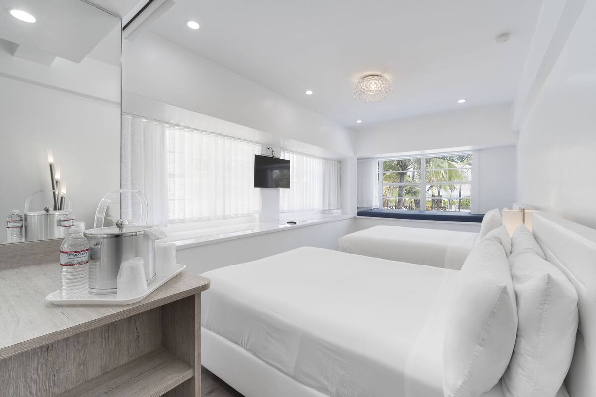 A hotel room with two beds, a large mirror, a TV mounted on the wall, and a window with a view of palm trees.