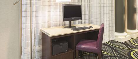Quarto de hotel com escrivaninha, computador, impressora e uma cadeira roxa.