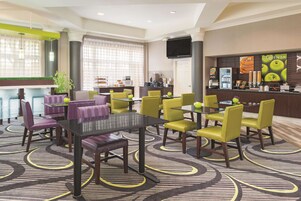 Un vestíbulo de hotel con mostrador de recepción, un área de comedor con sillas amarillas, una pantalla de televisión grande y una alfombra estampada.