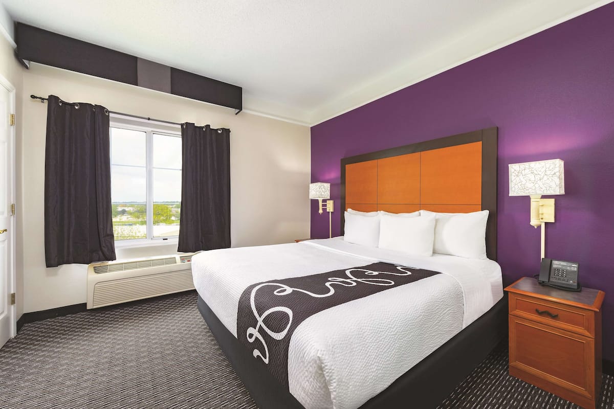 Une chambre d’hôtel avec un grand lit, une fenêtre avec des rideaux, un radiateur, une table de chevet avec une lampe et un mur de couleur violette.