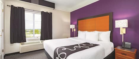 Una habitación de hotel con una cama grande, una ventana con cortinas, un calefactor, una mesita de noche con lámpara y una pared morada.