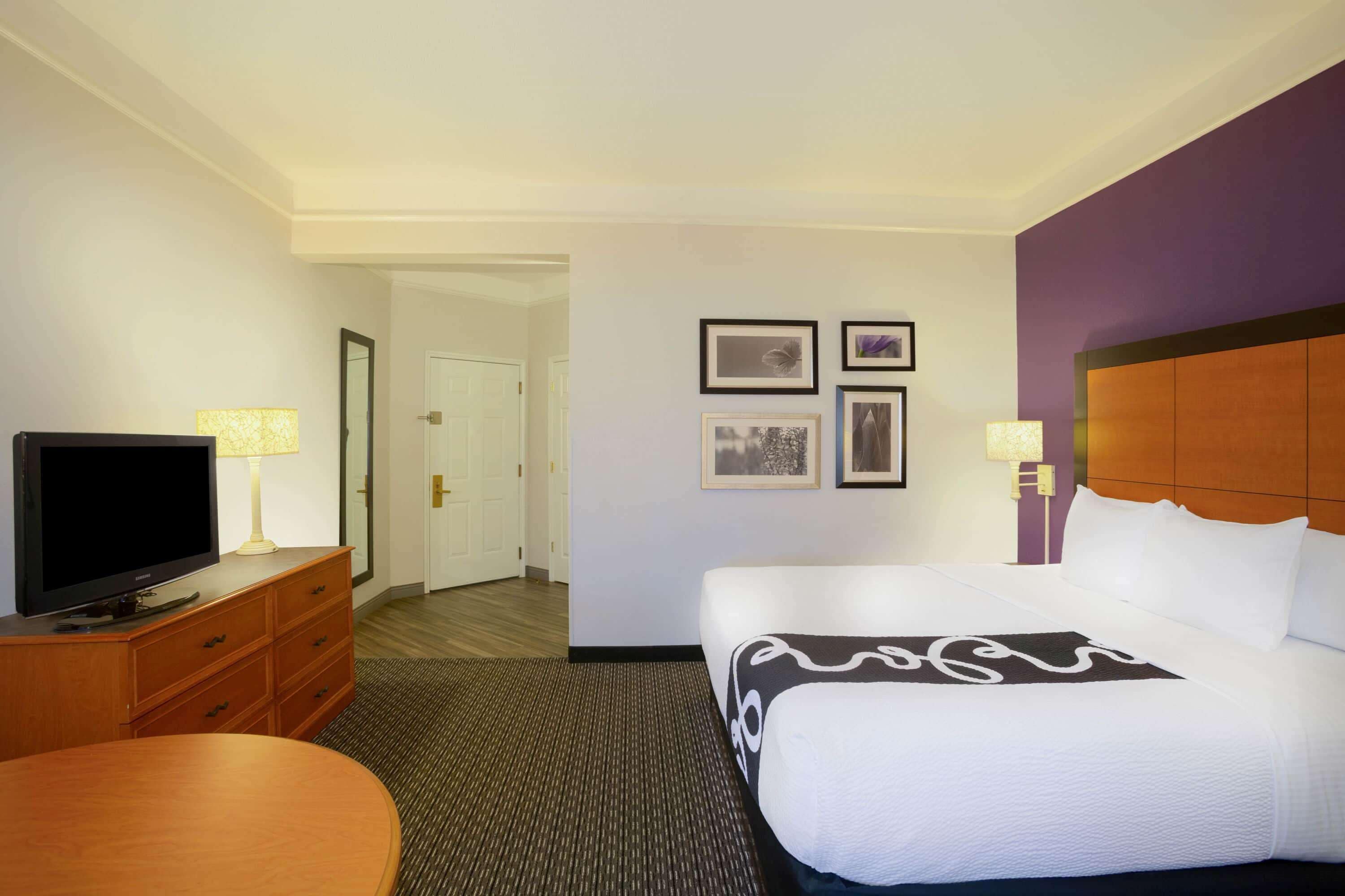 Une chambre d’hôtel avec un lit, un téléviseur posé sur une commode en bois, un bureau et une chaise. Des tableaux encadrés ornent le mur, et une porte donne accès à une autre pièce.