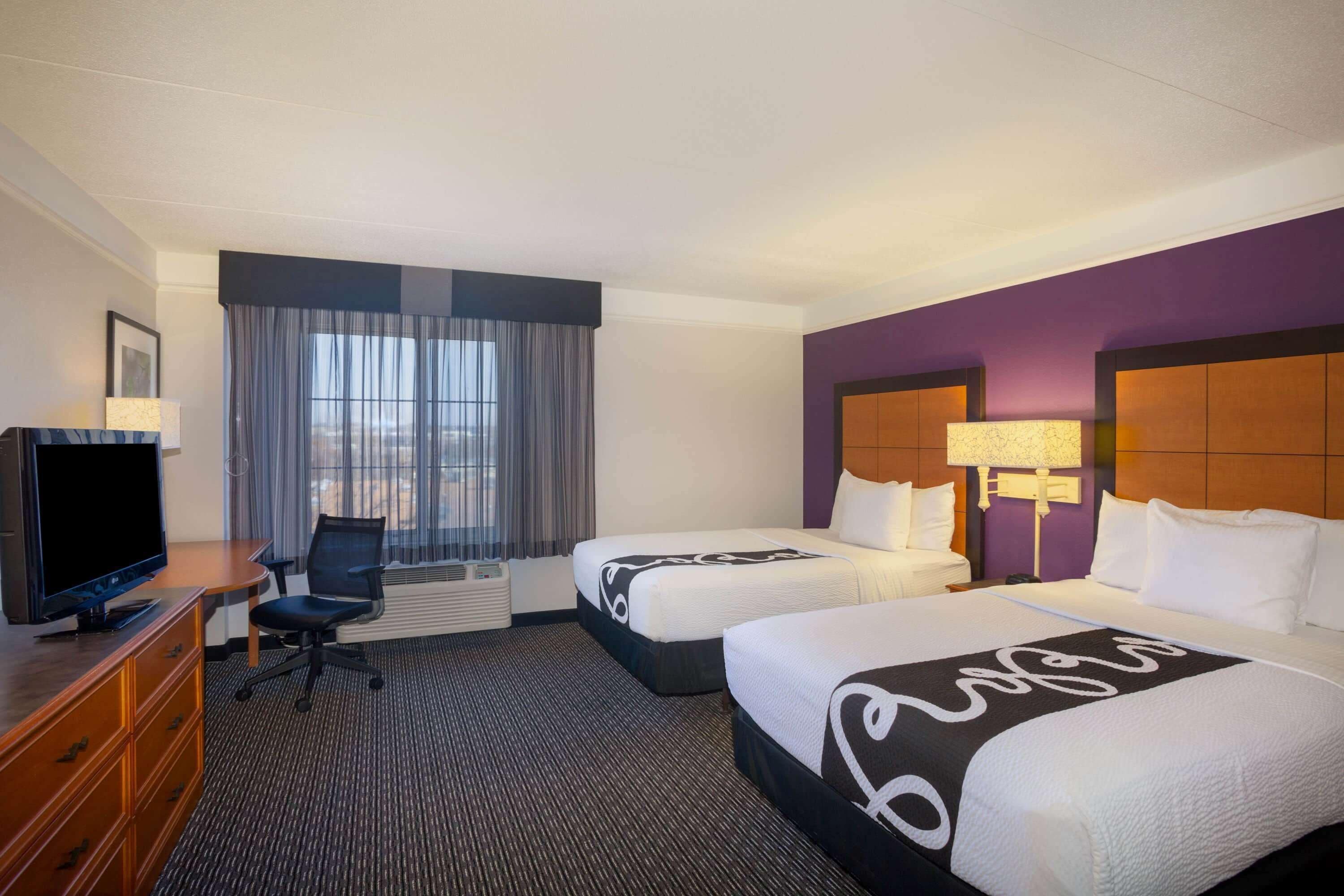 Quarto de hotel com duas camas, uma mesa de trabalho com televisão, uma cadeira e janela com cortinas.