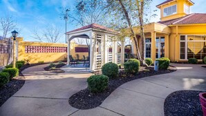 Un espace extérieur aménagé, abrité et doté d’une pergola, entouré d’arbustes décoratifs et bordé d’un chemin pavé.