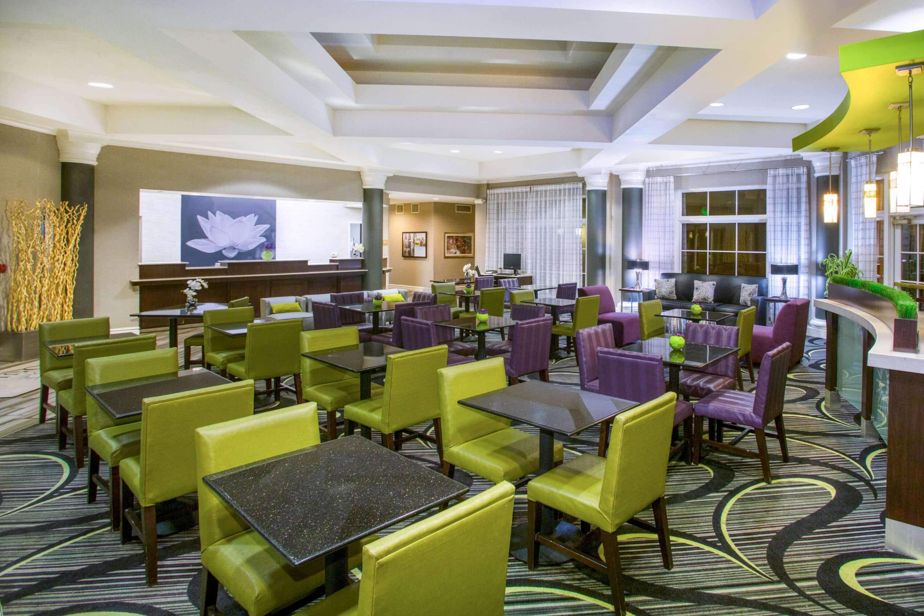 A hotel lobby with multiple seating areas, including tables and chairs, a reception desk, and a large wall-mounted screen.