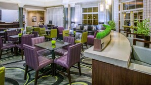 Un hall d’hôtel doté de plusieurs espaces aménagés avec des sièges, notamment des tables rondes et des canapés, ainsi qu’un comptoir de réception agrémenté d’une plante verte.