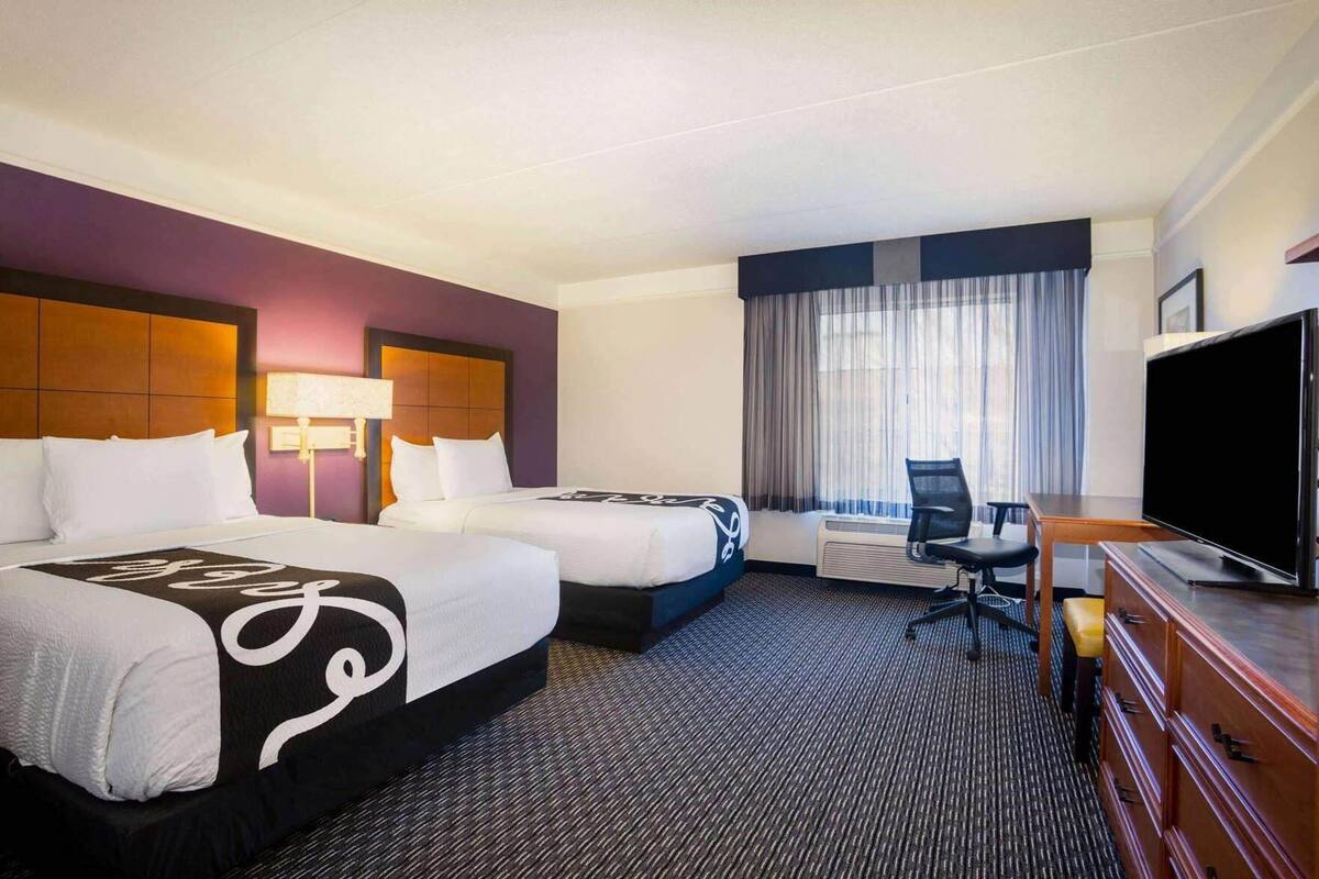 Une chambre d’hôtel avec deux lits, un bureau, une chaise, une télévision et une fenêtre avec des rideaux.