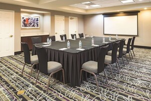 Un salón de conferencias con una larga mesa, sillas, botellas de agua y una pantalla de proyector.