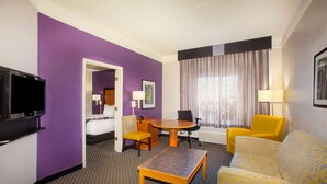 Une chambre d’hôtel avec des murs violets, un téléviseur à écran plat, un lit, une table à manger avec des chaises, un canapé et une fenêtre avec des rideaux.