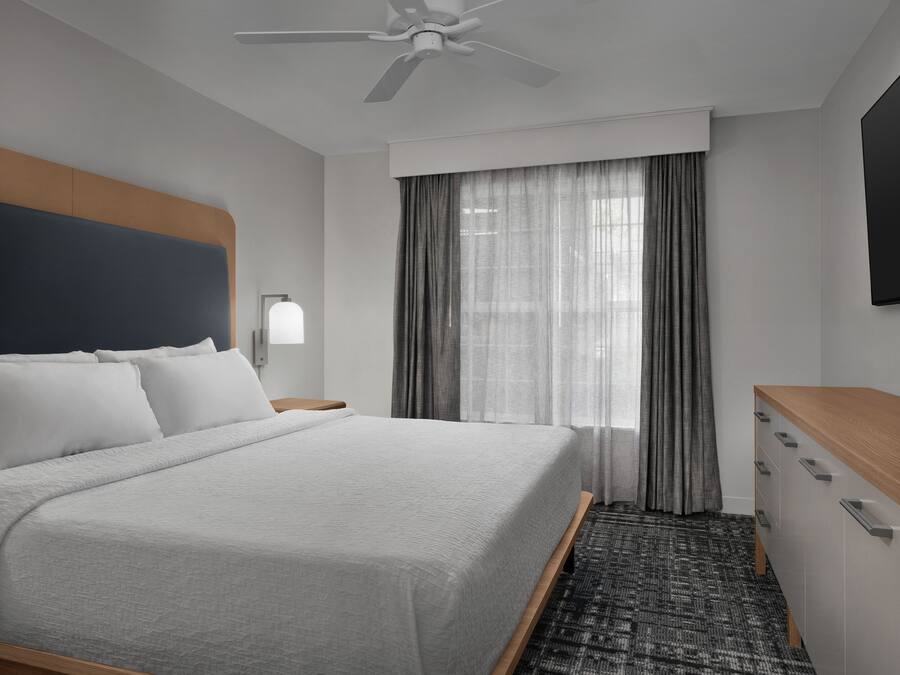 A hotel room with a bed, a desk, a flat-screen TV, a ceiling fan, and a window with curtains.