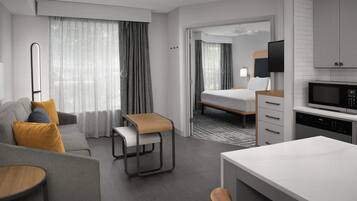 A modern hotel room with a sofa, a small table, a bed, a TV, a microwave, and a kitchen area.