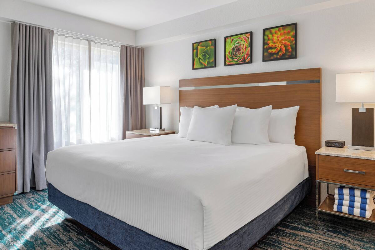 A hotel room with a large bed, two bedside tables, a nightstand, a lamp, and a wall-mounted clock.