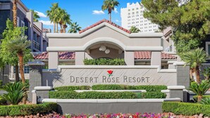 A sign for Desert Rose Resort with a red rose emblem, surrounded by landscaped greenery and palm trees. - Desert Rose Resort - No Resort Fee (Las Vegas)
