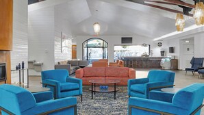 Un hall d’entrée spacieux, avec un grand canapé orange, des fauteuils bleus et une table basse. Au fond, on aperçoit un coin cuisine avec un bar et un téléviseur.
