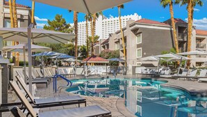 A pool area with lounge chairs and umbrellas, surrounded by palm trees and a building with balconies. - Desert Rose Resort - No Resort Fee (Las Vegas)
