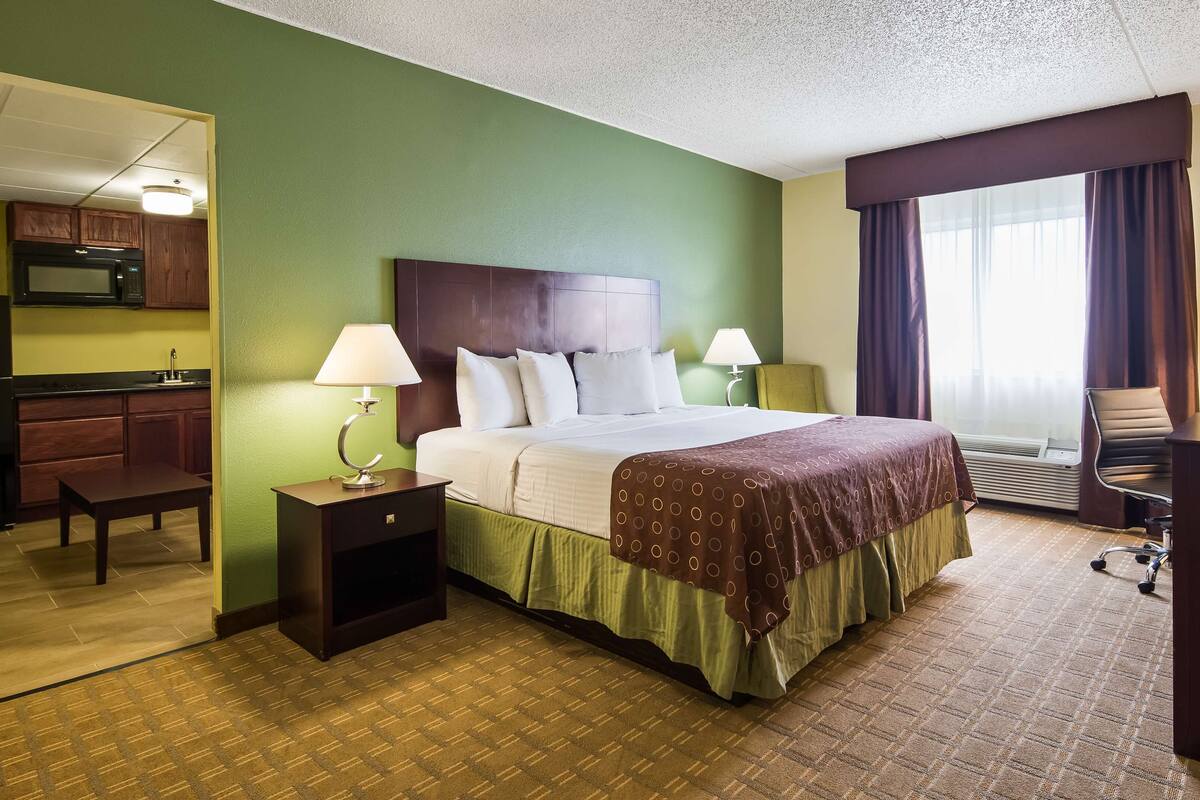 A hotel room with a bed, bedside lamps, a desk with a chair, a television, and a kitchenette.
