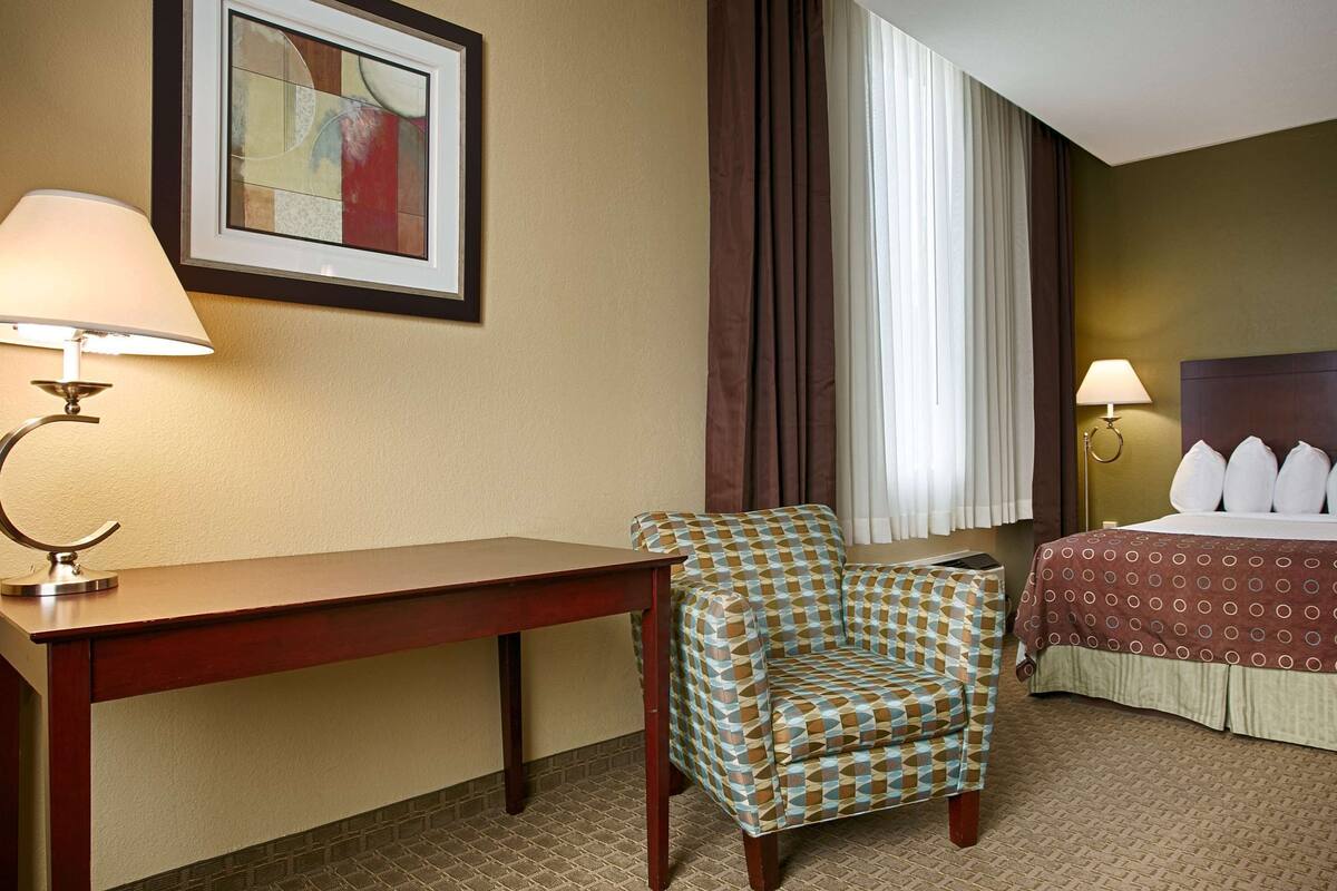 A hotel room with a bed, a desk, a chair, a lamp, a painting on the wall, and a window with curtains.
