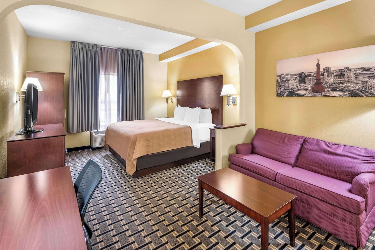 A hotel room with a bed, a sofa, a desk, a TV, and a painting on the wall.