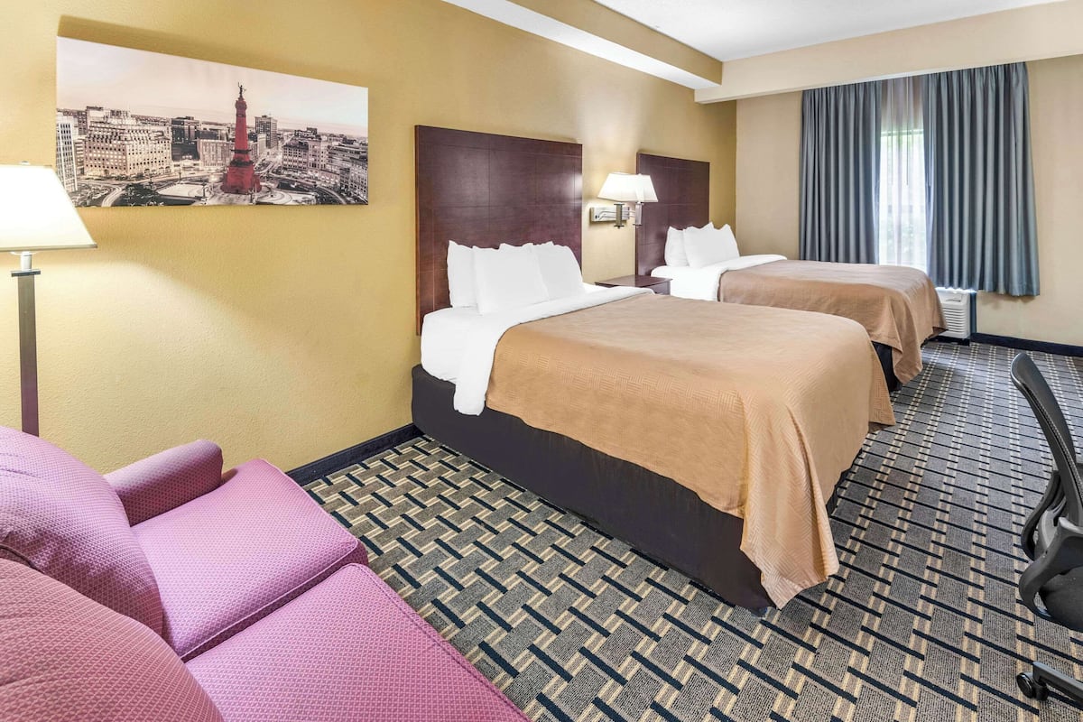 A hotel room with two beds, a sofa, a lamp, and a cityscape painting on the wall.