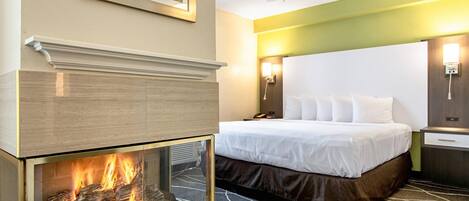 A hotel room with a fireplace, a bed with white bedding, a ceiling fan, and a nightstand with a lamp.