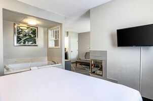 A modern hotel room with a large bed, a mounted TV, a fireplace, and a bathroom with a bathtub and shower.