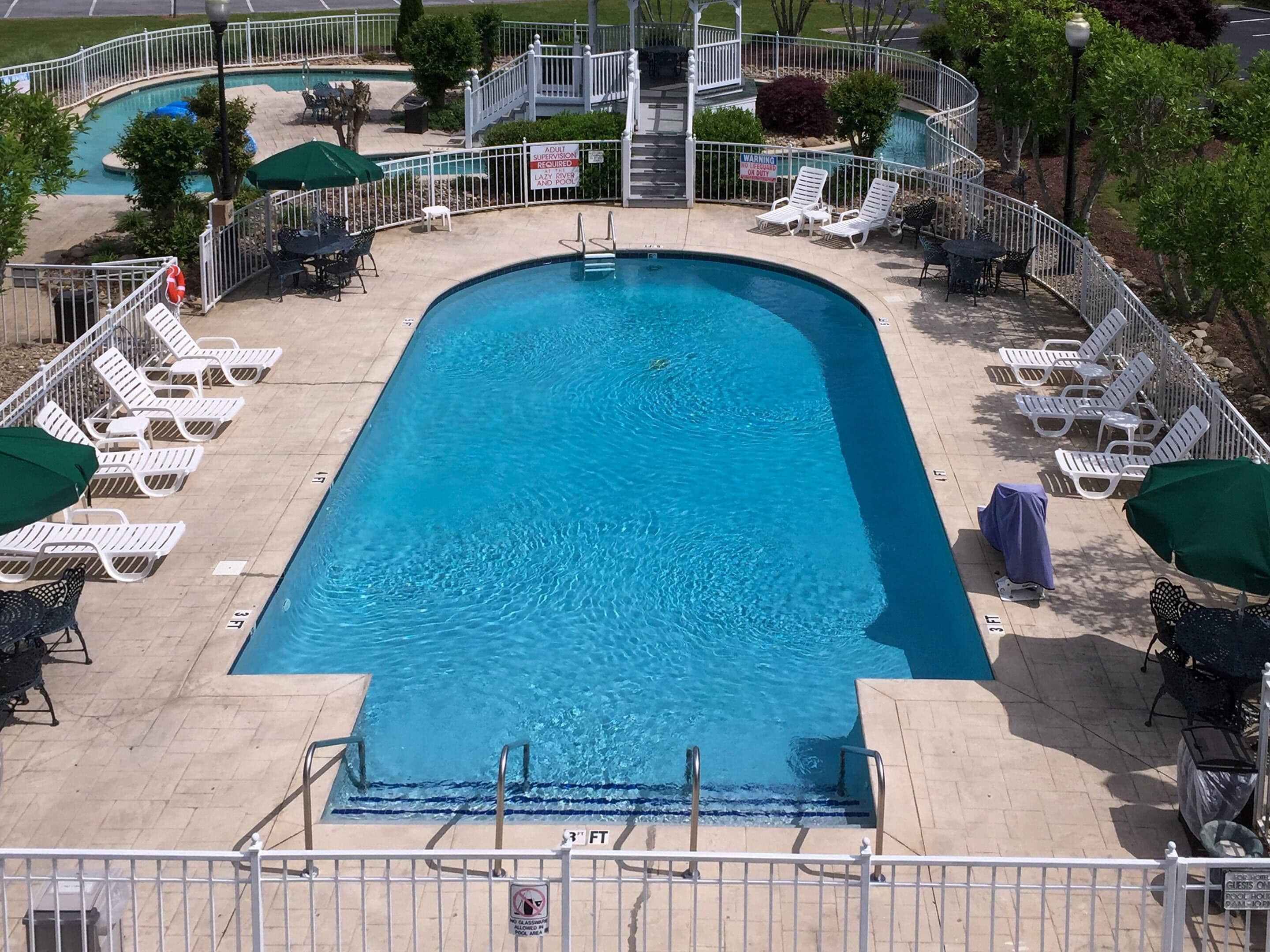 A swimming pool with lounge chairs and umbrellas, surrounded by a white fence and a covered seating area.