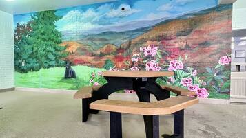 Una habitación con un mural de un paisaje, una mesa de madera con bancos y una pared de ladrillo.