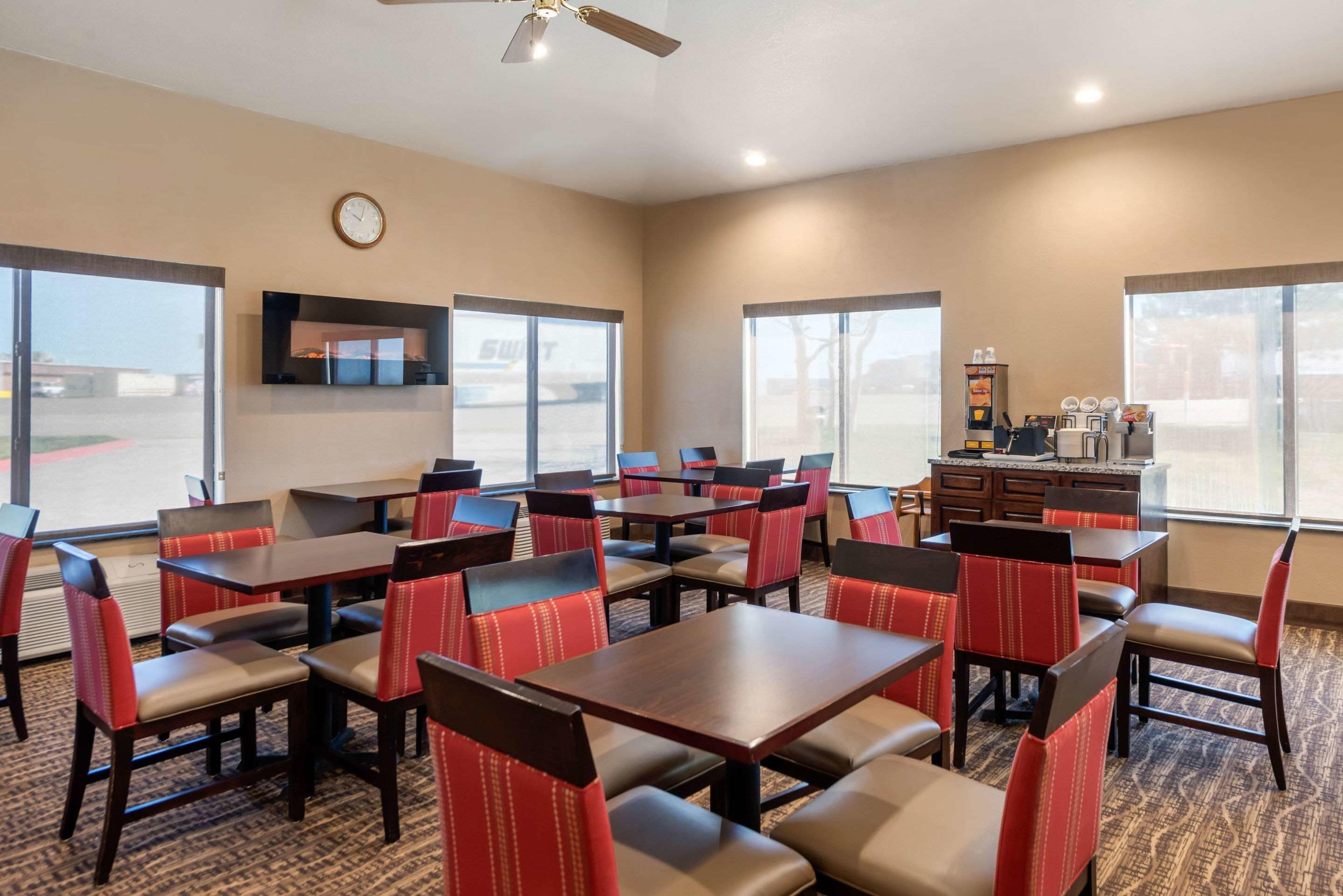 A hotel dining area with multiple tables and chairs, a clock on the wall, and a view of the parking lot through large windows.
