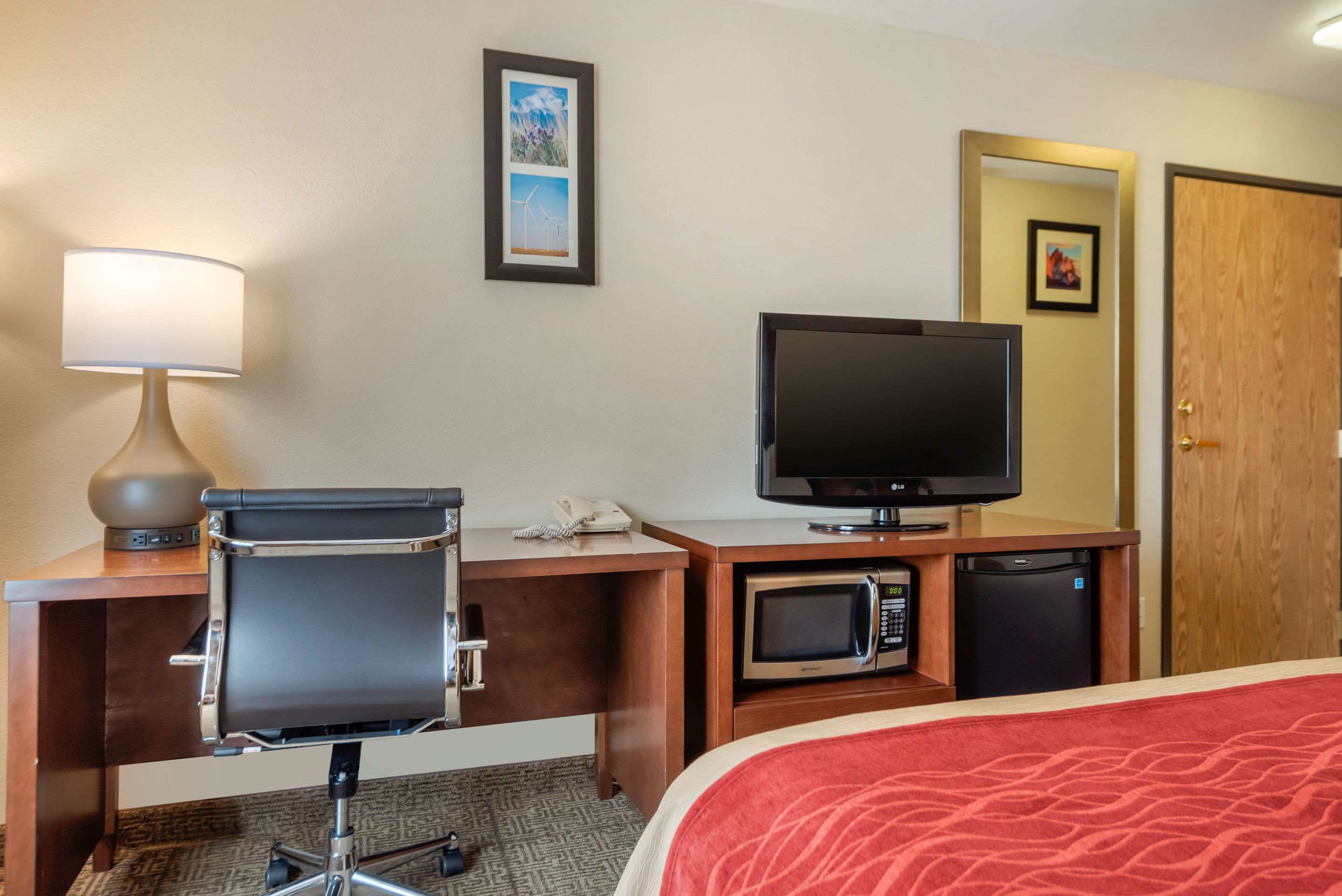 A hotel room with a desk, a chair, a television, a microwave, and a bed with a red blanket.