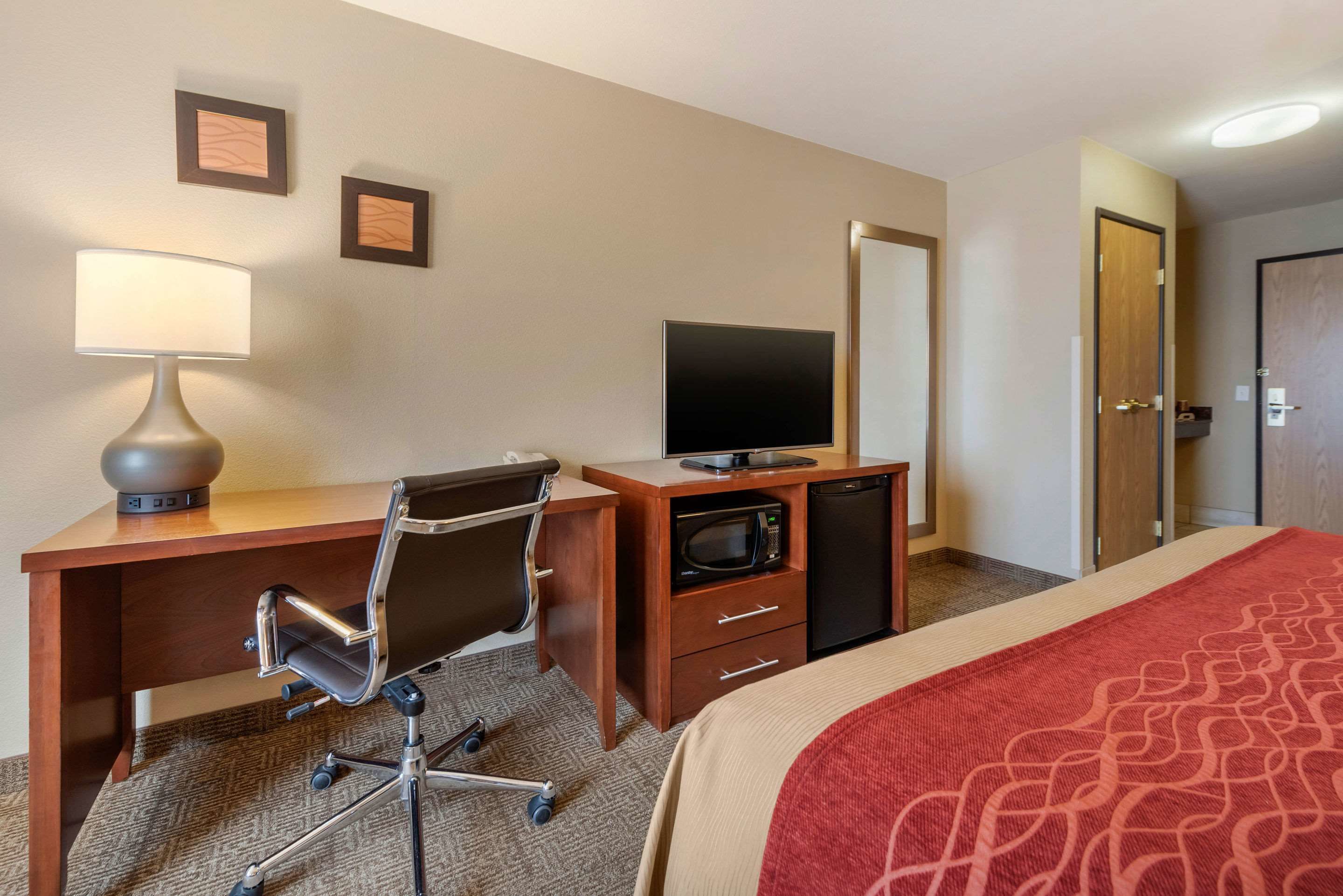 Habitación de hotel con escritorio, silla, televisor, microondas y cama con una colcha roja.