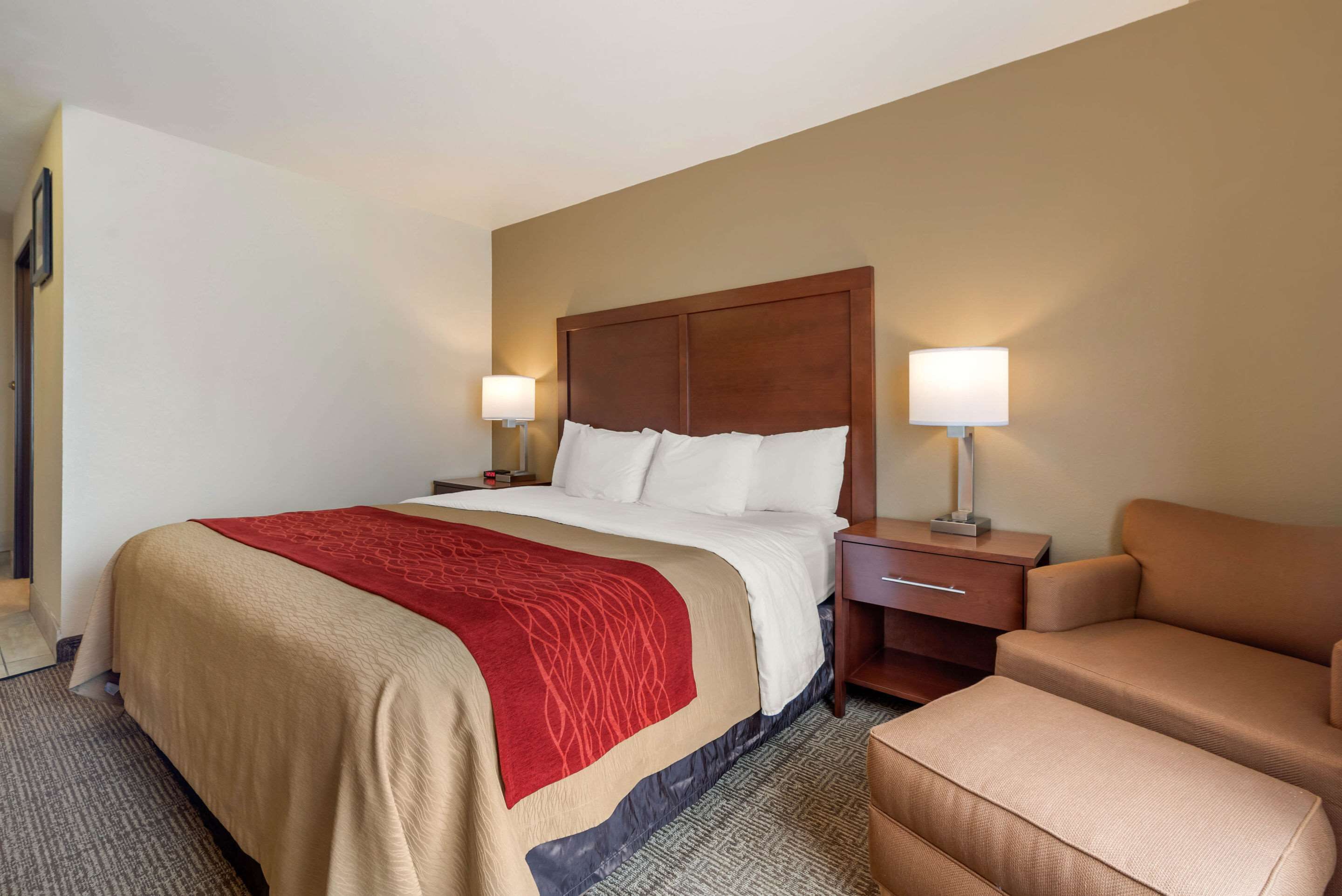A hotel room with a large bed, two bedside tables with lamps, a red throw blanket, and a brown sofa.