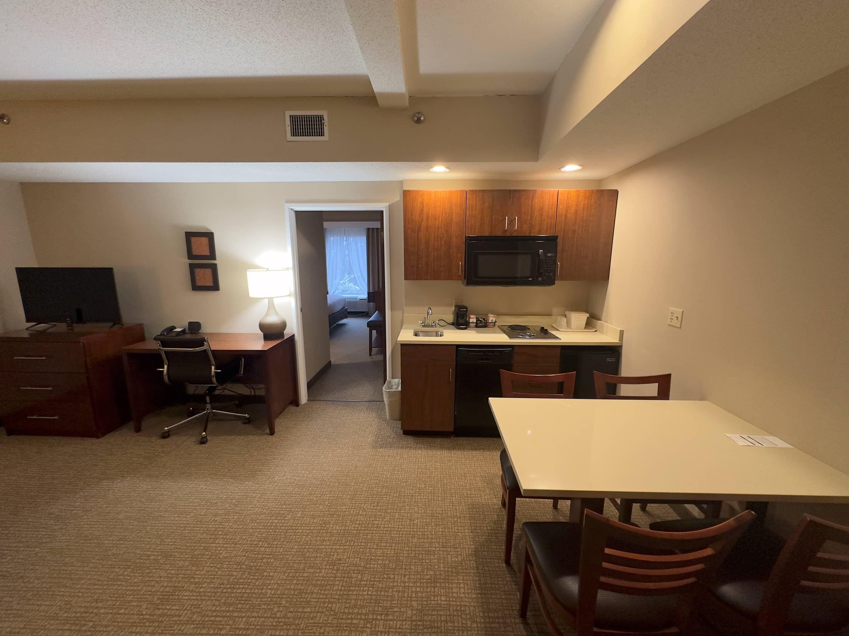 A hotel room with a kitchenette, a dining area, a desk with a chair, and a TV.