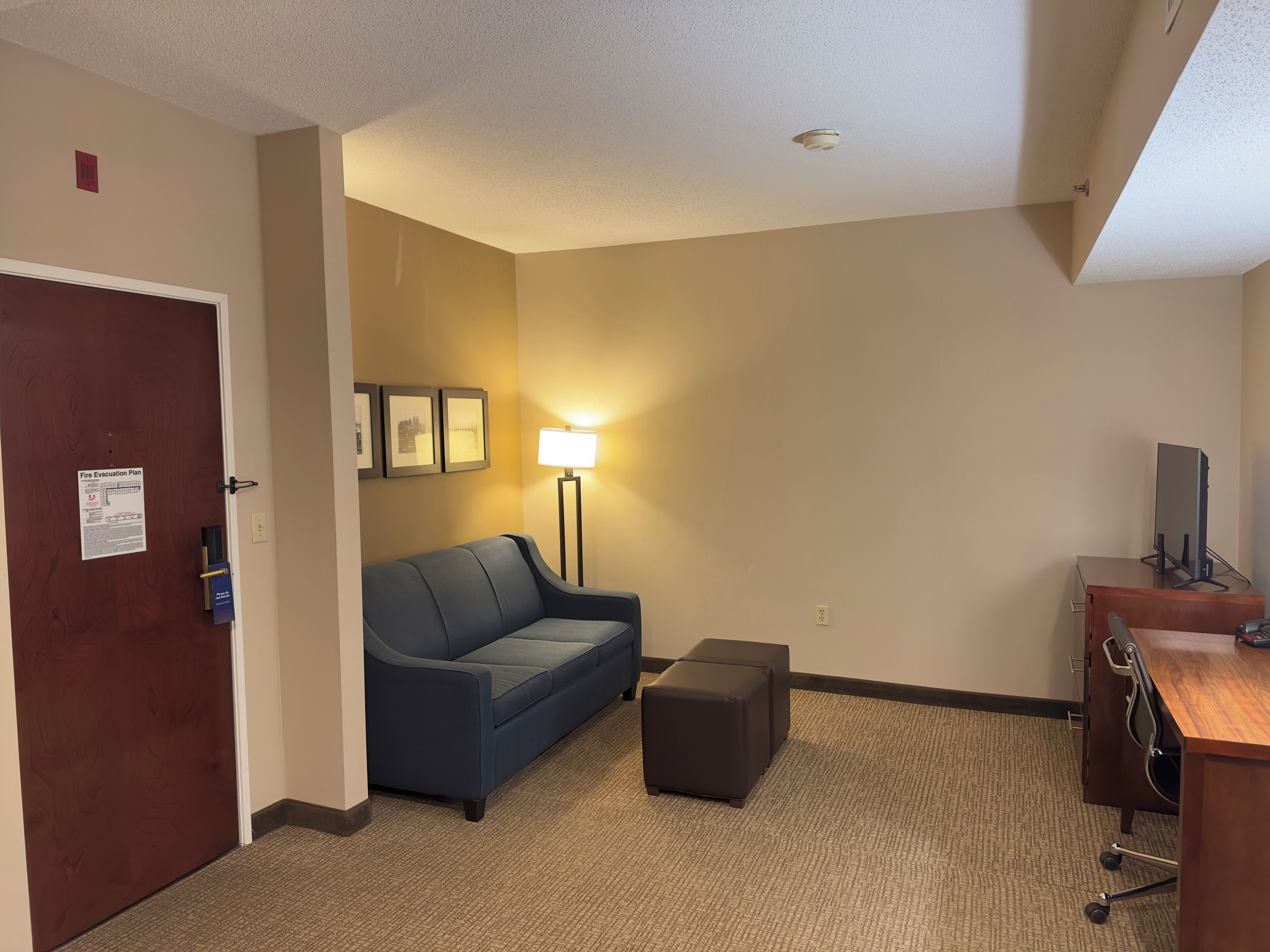A hotel room with a wooden door, a blue sofa, a standing lamp, a small ottoman, and a desk with a computer.