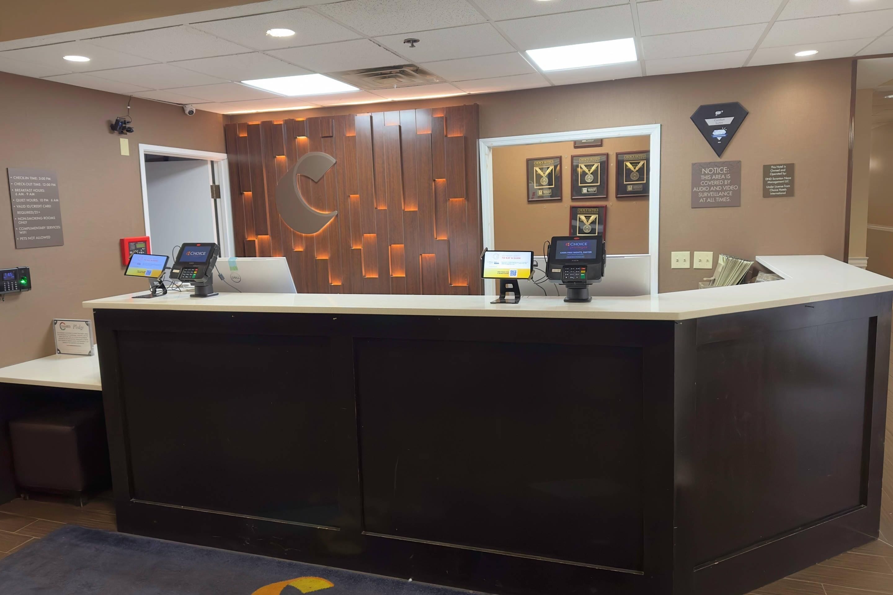 Un comptoir d’accueil avec un panneau décoratif en bois, un kiosque numérique et des trophées encadrés accrochés au mur.