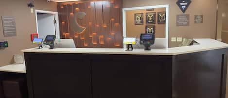 Un comptoir d’accueil avec un panneau décoratif en bois, un kiosque numérique et des trophées encadrés accrochés au mur.