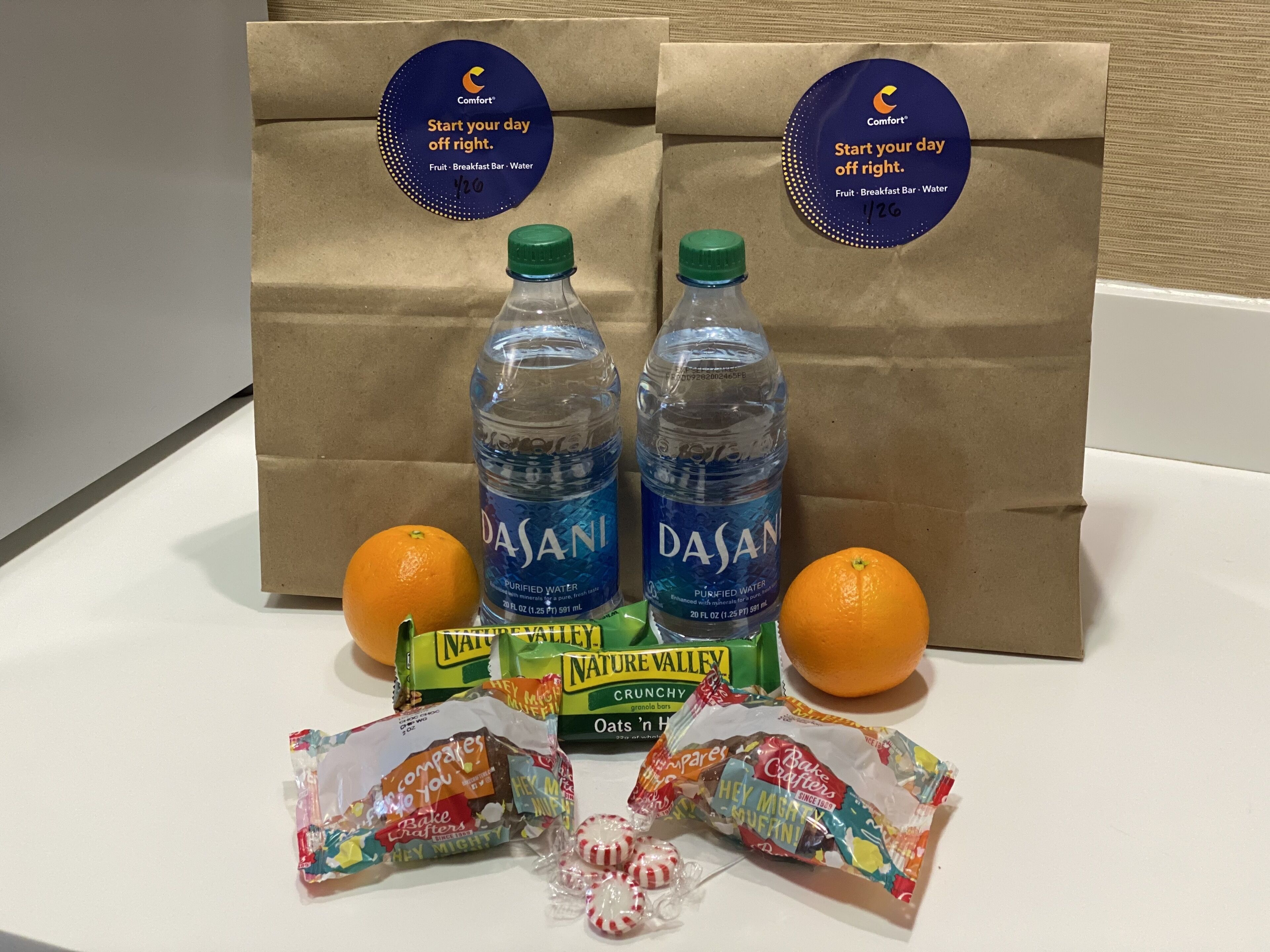Uma mesa com duas garrafas de água Dasani, duas laranjas e vários pacotes de lanches, incluindo Nature Valley Crunchy Oats e Bake Cafters.