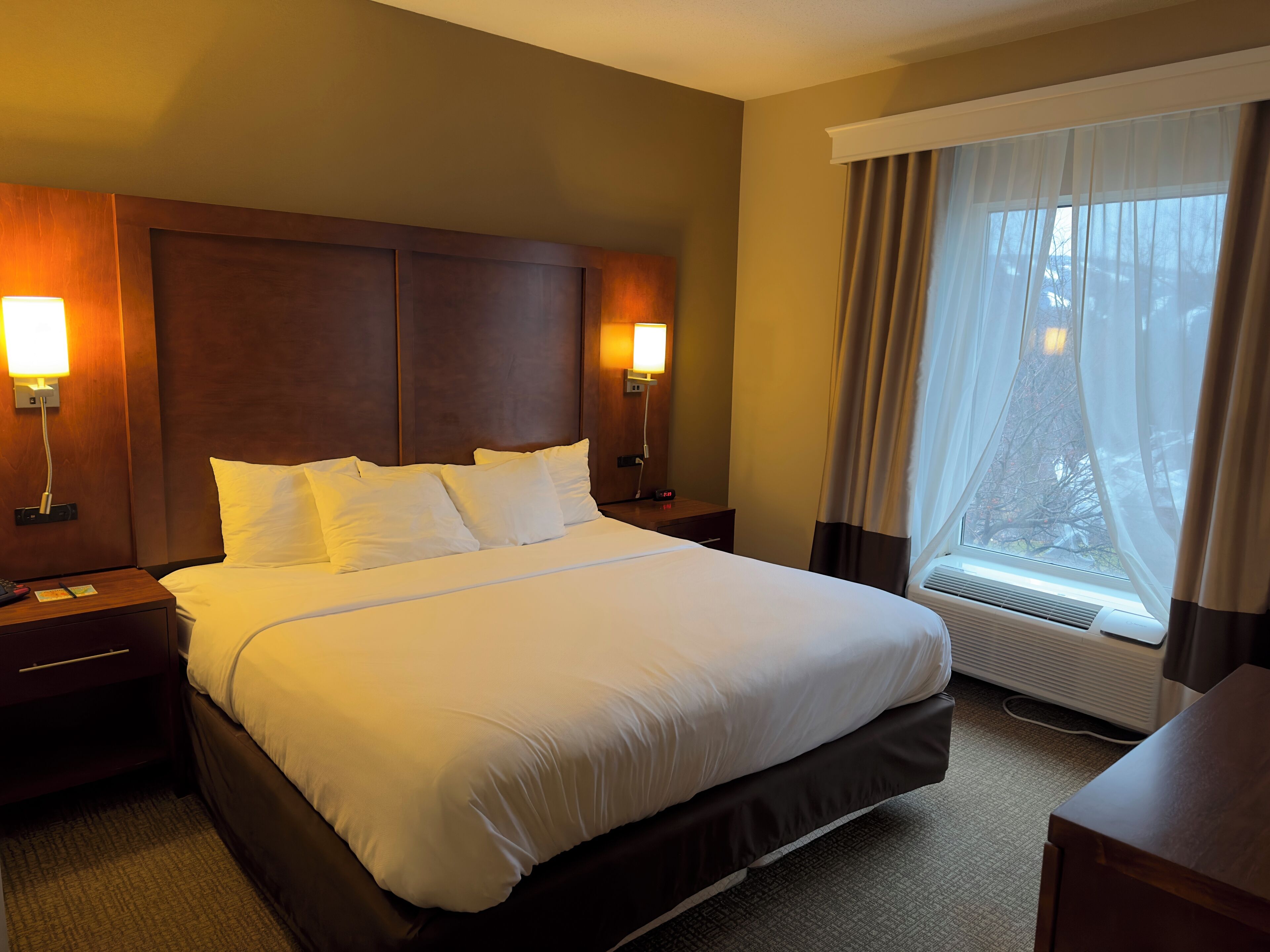 Habitación de hotel con una cama grande, dos mesitas de noche con lámparas, un ventanal con cortinas translúcidas y un equipo de aire acondicionado integrado.