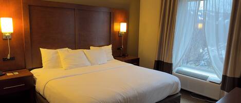 A hotel room with a large bed, two bedside tables with lamps, a window with sheer curtains, and a built-in air conditioning unit.