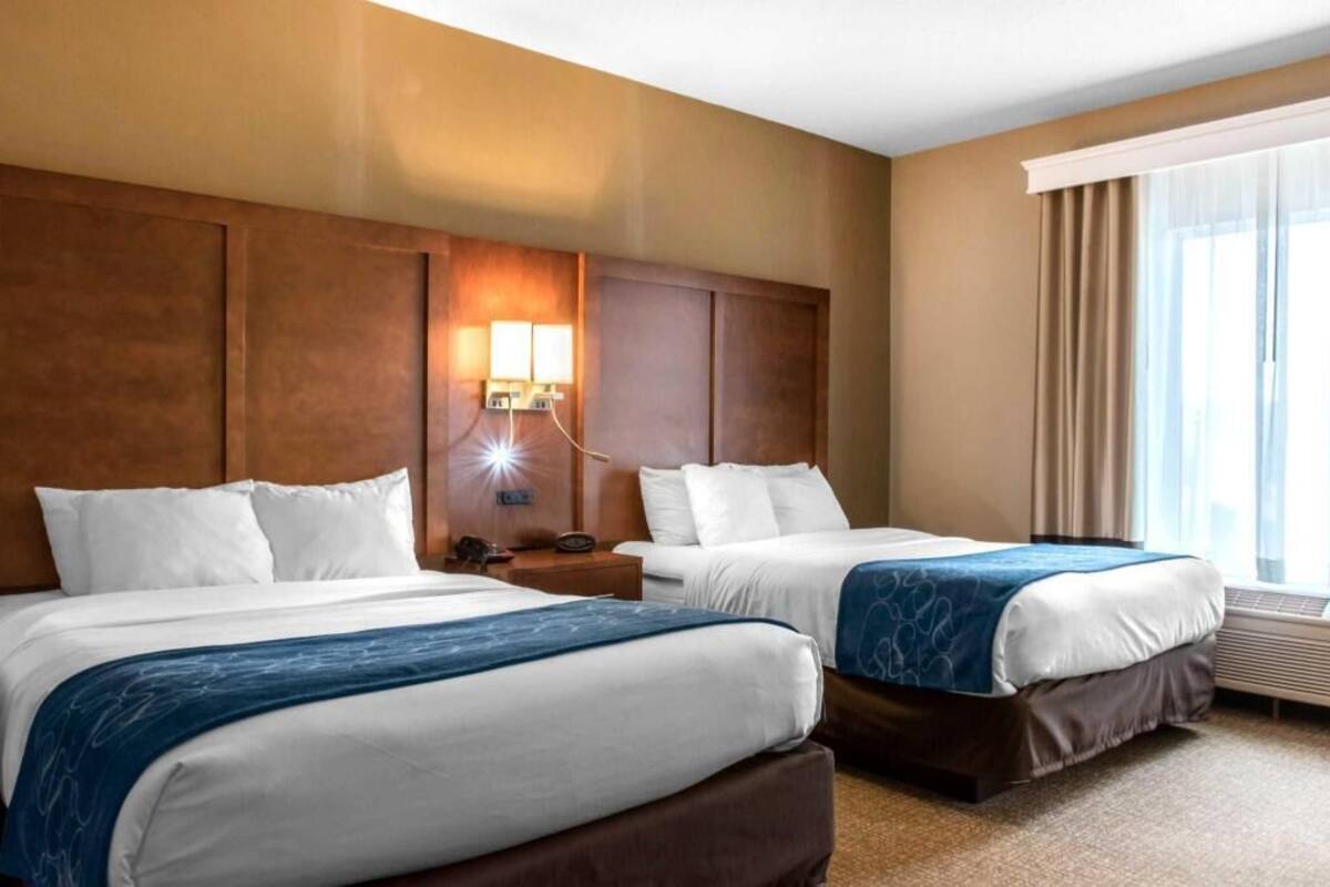 A hotel room with two beds, a nightstand with a lamp, a window with curtains, and a wall-mounted heater.
