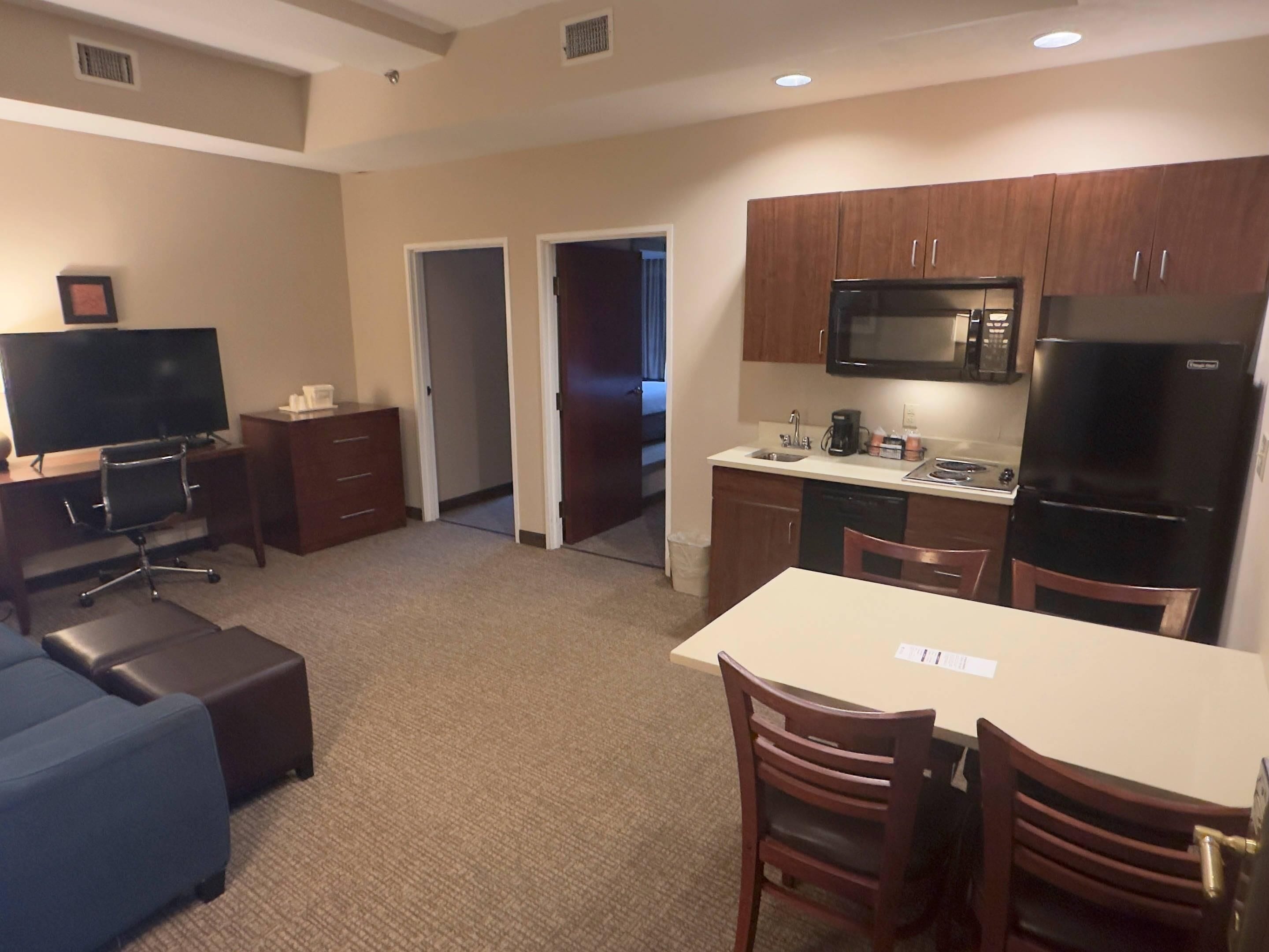 A compact hotel room with a kitchenette, a dining area with a table and chairs, a TV, and a sofa.
