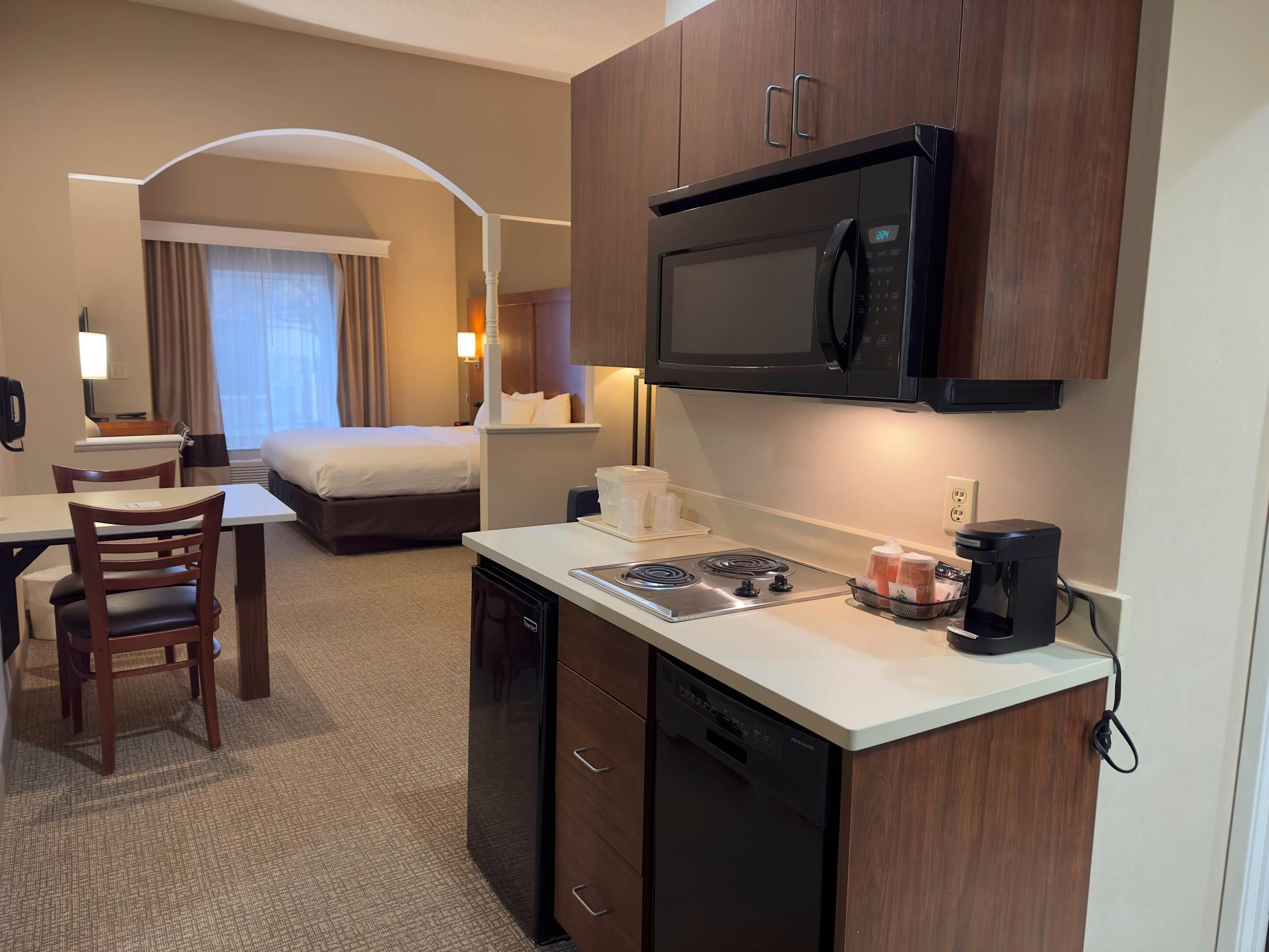 A hotel room with a kitchenette, including a microwave, stove, and coffee maker. There is a bed, a desk, and a chair.